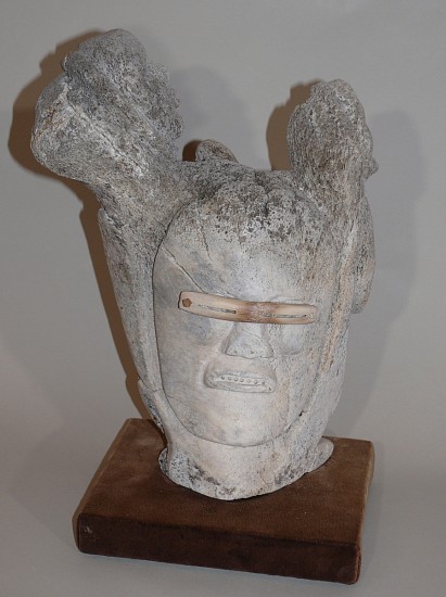 Surulak, Head with goggles and helping spirits, c. 1970
15 1/2 x 11 1/2 x 7 in.
A powerful whalebone head wearing snow goggles, with two helping spirits.  The sensitive and well-delineated face, accented with the antler snow goggles, contrasts with the two helping spirits, with just the barest suggestions of faces.  This is afine example of the limitations that whalebone as a medium imposed on artists.  Where (as on the helping spirits) the outer surface of the bone had been worn away, carving was difficult and perilous.
03707-1