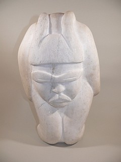 Osuitok Ipeelee, Kakagun, the Great Hunter, c. 1970
16 x 10 x 6 in.
A massive, enigmatic portrait by Osuitok Ipeelee, one of the premier first-generation Inuit artists.  The sensitive face sports snow goggles, and a puzzling pair of horns extends over the figure's forehead.  The rest of the sculpture is largely without details.

Osuitok Ipeelee was one of the most talented, versatile, and prolific Inuit sculptors.  His subjects ranged from people to animals to transformations.  His style varied from hyper-realistic to geometric to abstract.  With the exception of a series of strikingly beautiful caribou, he almost never repeated himself.

Most of Osuitok's works are in serpentine, which is softer and takes details more easily than the white Andrew Gordon Bay marble.  Even so, the horns have a delicate spiral texture, and the man's moustache is clearly textured.  Cape Dorset artists generally preferred serpentine to marble.  In "Northern Rock," Osuitok is quoted as saying, "I wasn't the only one working with the white stone from Andrew Gordon Bay back then [in the early 1970s].  Pauta Saila did too.  We used that marble in the winter because those stones were the only ones available in the hamlet."
03529-1
